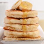 Healthy Greek yogurt pancakes stacked and topped with sliced banana and a drizzle of maple syrup