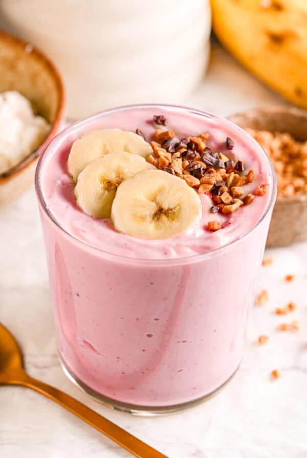 A creamy strawberry cottage cheese smoothie in a glass, topped with banana slices and crunchy cacao nibs.