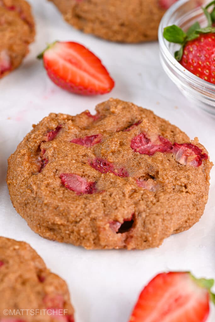 Close-up of freshly baked protein Greek yogurt cookies made with Greek yogurt and diced strawberries. The cookie has a soft texture with juicy strawberry pieces visible throughout. A sliced strawberry is placed beside it on parchment paper.