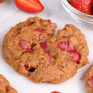 A Greek yogurt protein cookie with a cracked center, revealing its moist interior with vibrant red strawberry chunks. The cookie rests on parchment paper, surrounded by more cookies and fresh strawberries in a glass bowl.