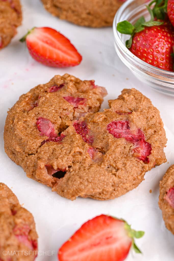 A Greek yogurt protein cookie with a cracked center, revealing its moist interior with vibrant red strawberry chunks. The cookie rests on parchment paper, surrounded by more cookies and fresh strawberries in a glass bowl.