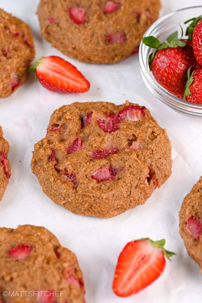 A batch of Greek yogurt protein cookies with diced strawberries, laid out on parchment paper. The cookies have a golden-brown surface with bright red strawberry pieces, and a glass bowl of fresh strawberries is placed beside them.