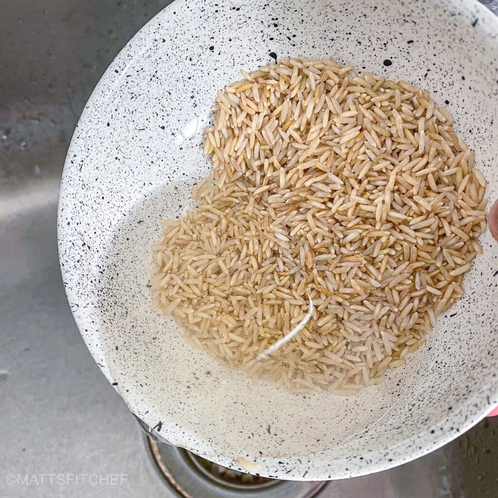 Rinsing brown rice in a speckled bowl under running water.