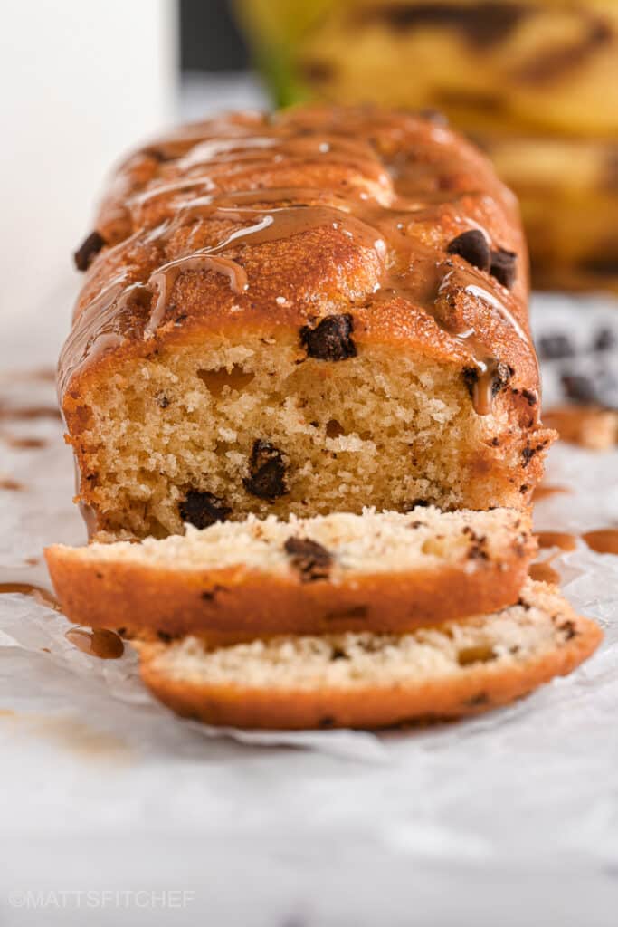 Close-up of sliced peanut butter banana bread loaf with chocolate chips, drizzled with peanut butter on top, sitting on parchment paper. The golden-brown crust and moist interior with melted chocolate chips are visible.