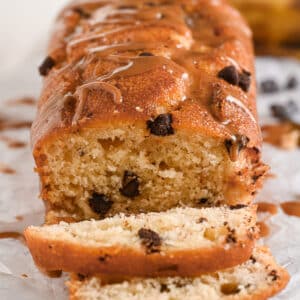 A freshly baked peanut butter banana bread loaf with golden-brown edges, chocolate chips, and a glossy peanut butter drizzle. The soft texture is visible as a few slices are cut, showing the inside.