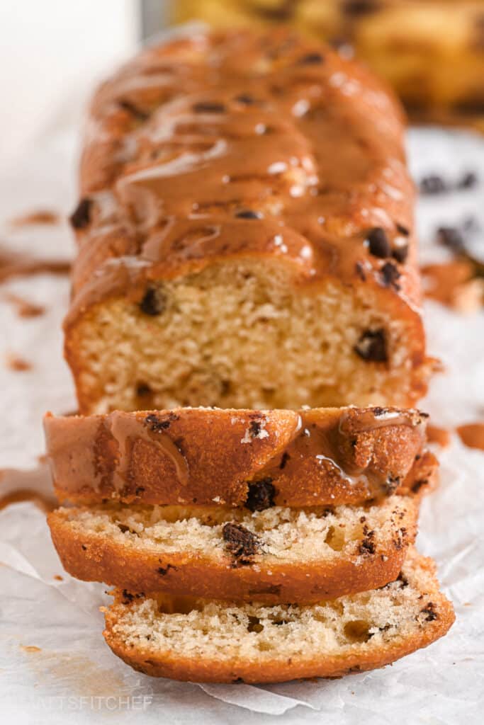 A stack of three slices of peanut butter banana bread with chocolate chips and a peanut butter drizzle on top. The slices rest on parchment paper, with crumbs and chocolate pieces scattered around.