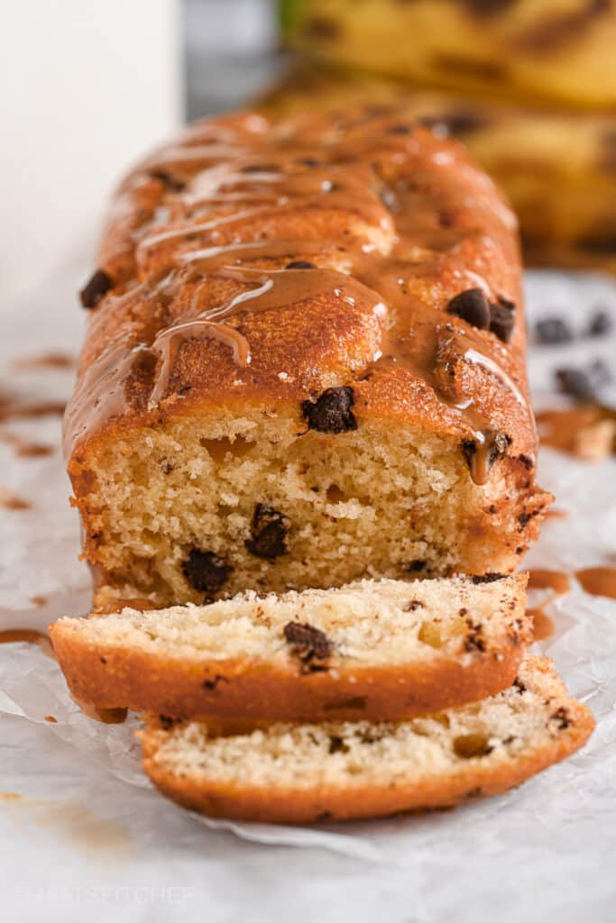 A freshly baked peanut butter banana bread loaf with golden-brown edges, chocolate chips, and a glossy peanut butter drizzle. The soft texture is visible as a few slices are cut, showing the inside.