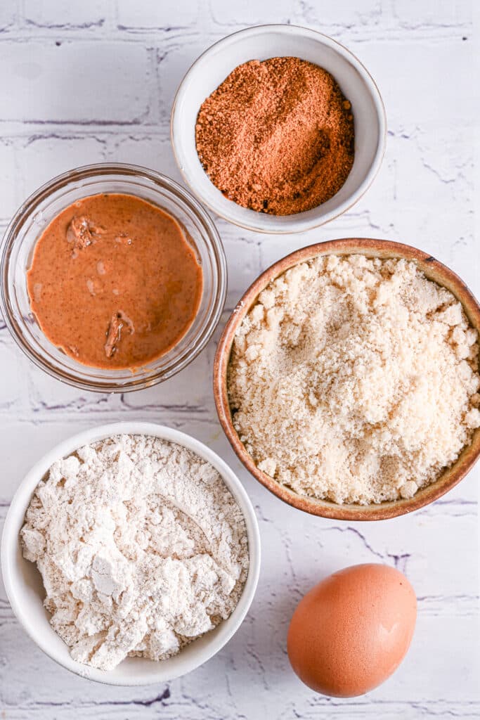 Almond flour, protein powder, peanut butter, and eggs displayed in bowls, showing all the essential ingredients for making homemade protein cookies