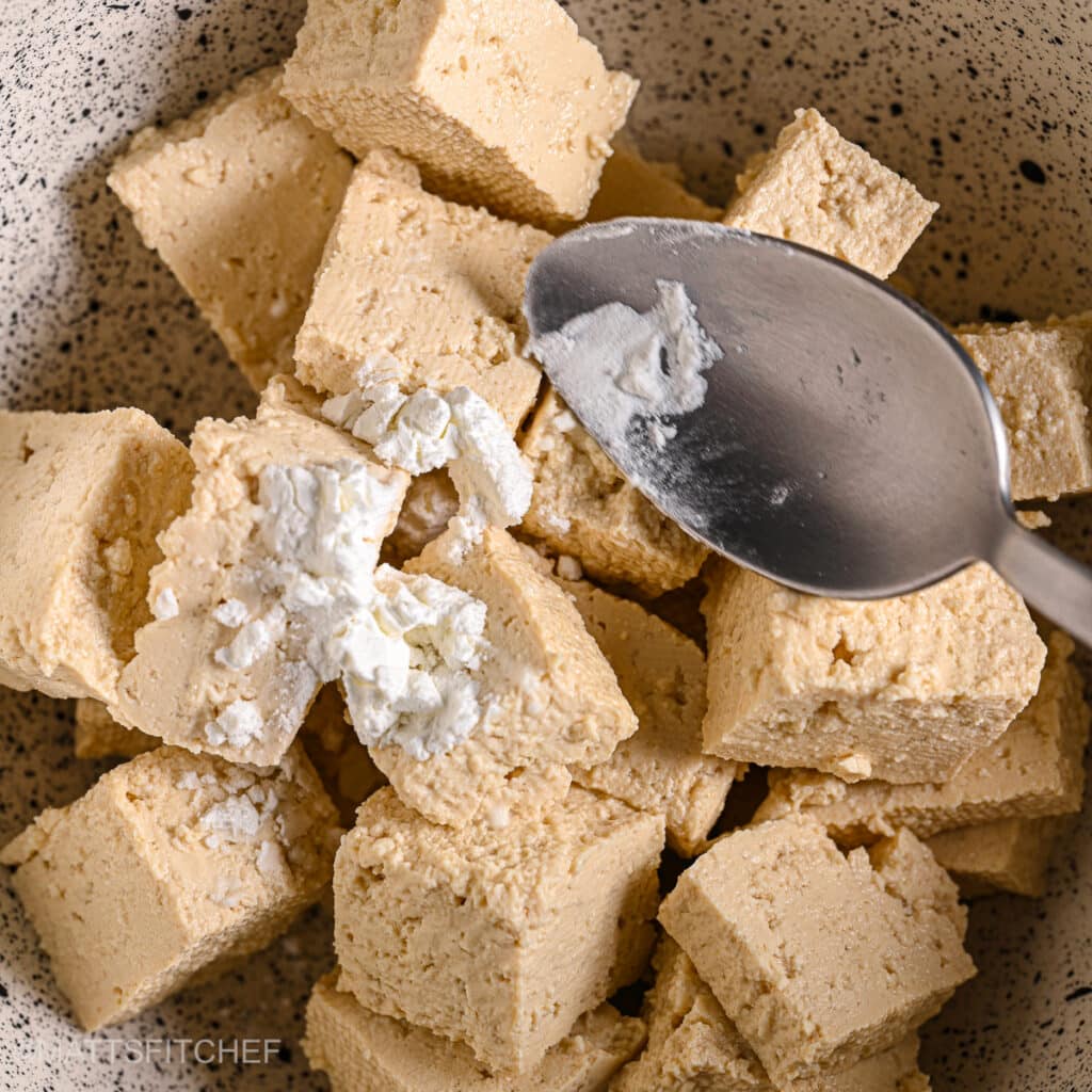 Extra-firm tofu cubes in a bowl being sprinkled with cornstarch for extra crispiness.