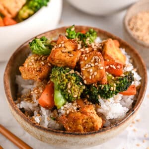 A bowl of crispy tofu stir fry with broccoli, carrots, and sesame seeds served over fluffy white rice, with chopsticks on the side.