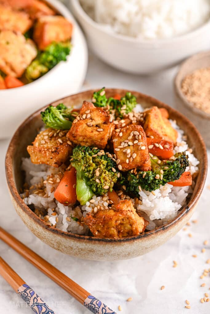 A bowl of crispy tofu stir fry with broccoli, carrots, and sesame seeds served over fluffy white rice, with chopsticks on the side.