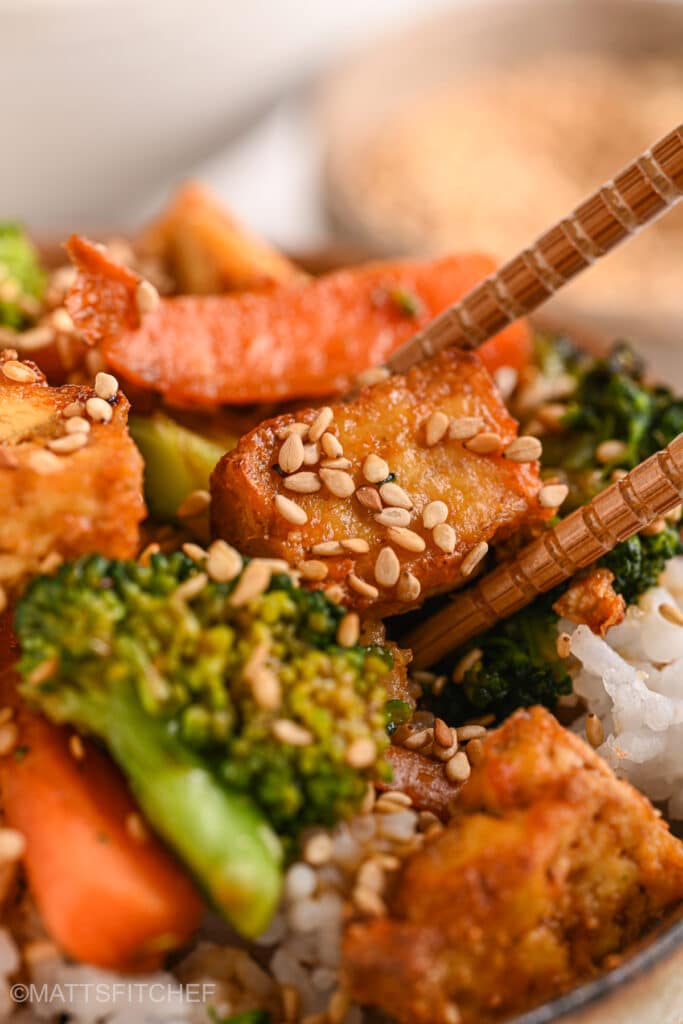 A pair of chopsticks picking up a flavorful piece of tofu from a bowl of stir fry.