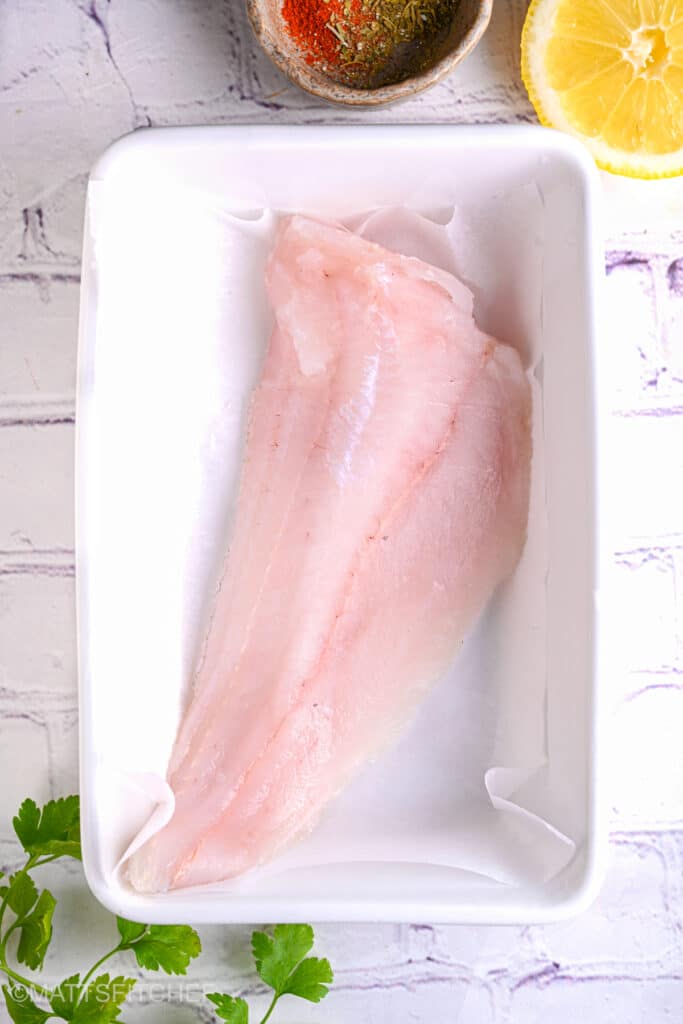 Raw cod fillet in a baking dish – Fresh cod fillet placed in a white baking dish lined with parchment paper, ready for seasoning.