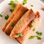 Overhead shot of oven baked crispy taquitos on a white plate, topped with fresh parsley for garnish.