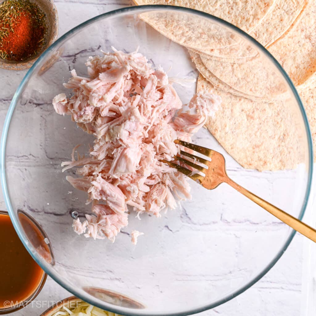 Shredding cooked chicken breast in a large glass bowl with a fork for chicken taquitos filling.