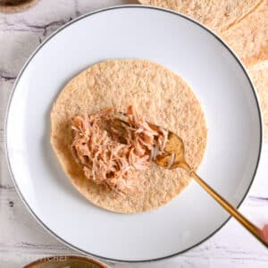 Spooning shredded chicken filling onto a tortilla to roll homemade chicken taquitos.