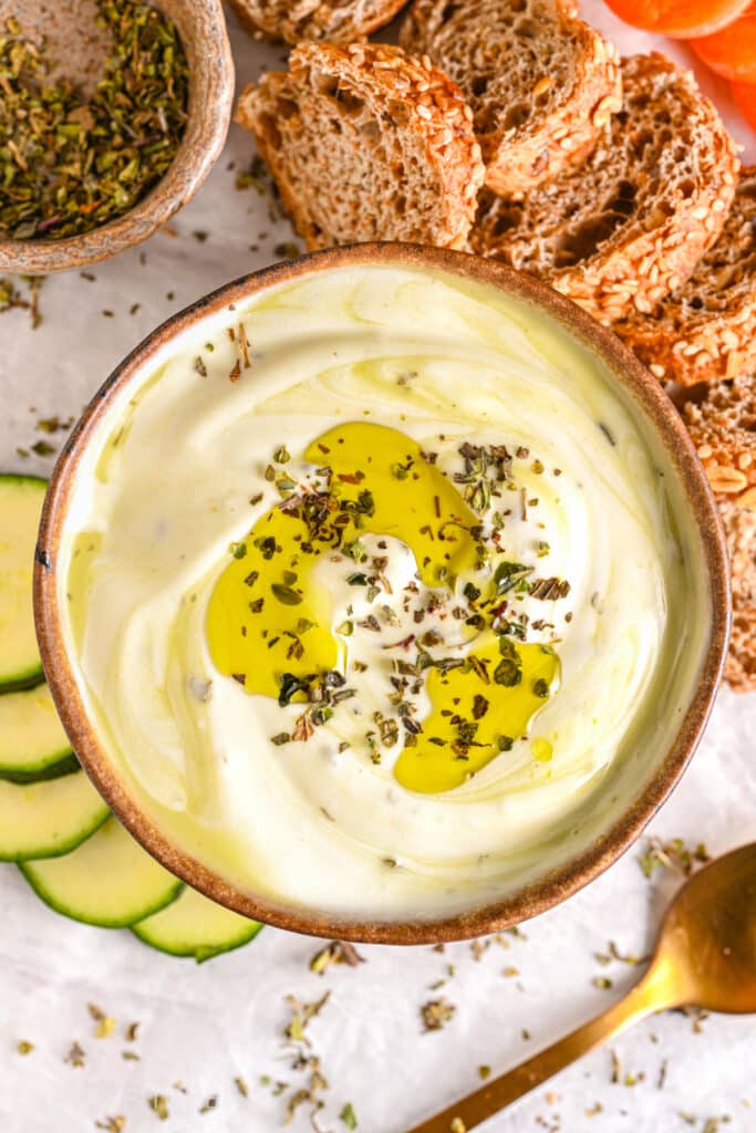 Creamy cottage cheese dip with a drizzle of olive oil and dried herbs, served with whole-grain bread, vegetables, and fresh zucchini slices.