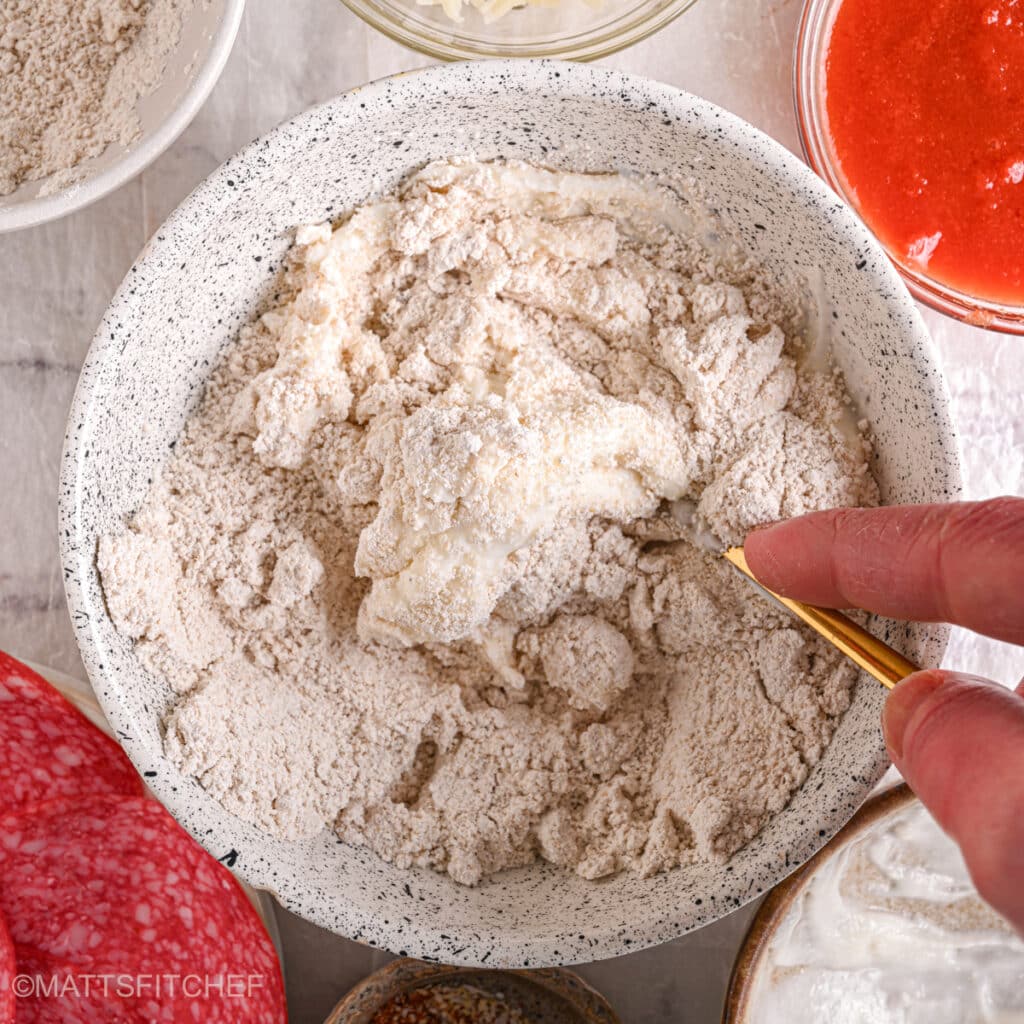 Mixing cottage cheese and flour – A hand stirring cottage cheese and flour mixture to form a macro-friendly pizza dough.