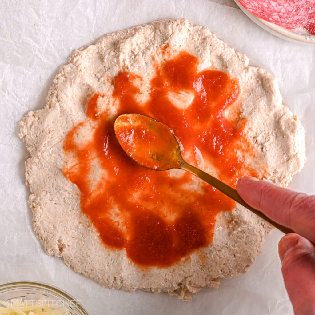 Spreading sugar-free pizza sauce – A spoon spreading homemade sugar-free pizza sauce over the high-protein pizza crust.