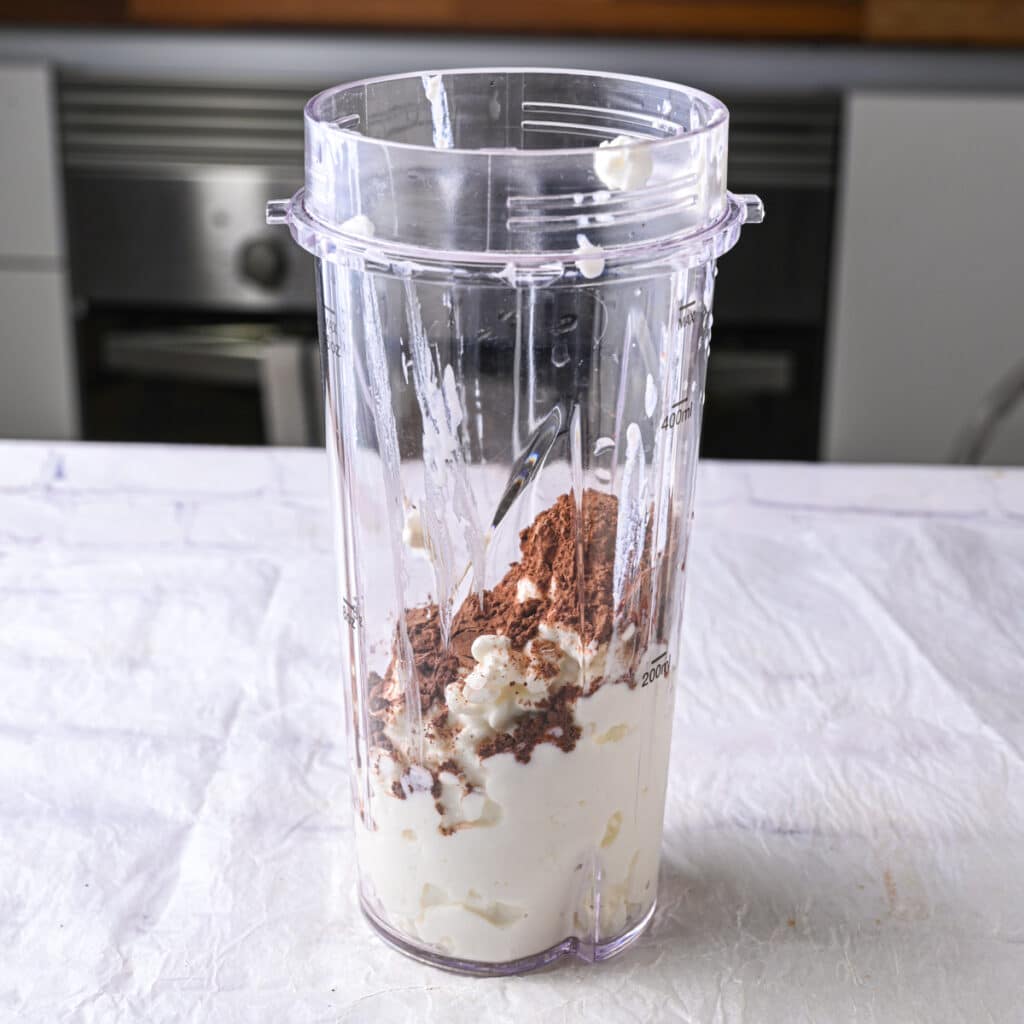 Blender cup filled with cottage cheese and cocoa powder, ready to be blended for homemade high-protein chocolate dessert.