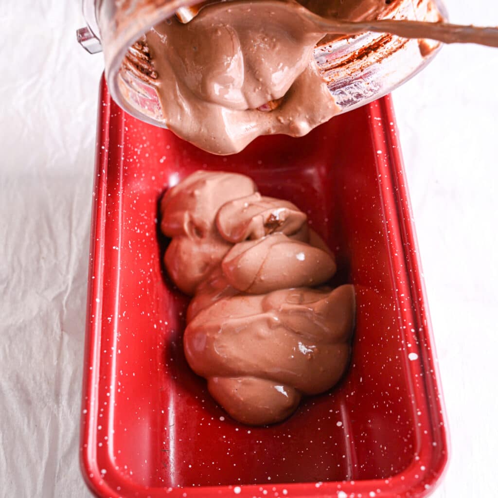 Thick and creamy chocolate cottage cheese ice cream being poured from a blender into a red loaf pan.