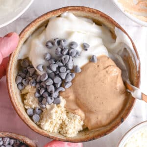 A golden spoon beginning to mix protein powder, peanut butter, Greek yogurt, and chocolate chips.