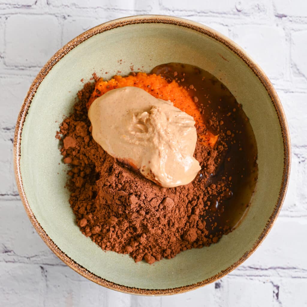Combining sweet potato brownie ingredients in a bowl &ndash; mashed sweet potato, almond butter, maple syrup and cocoa powder.