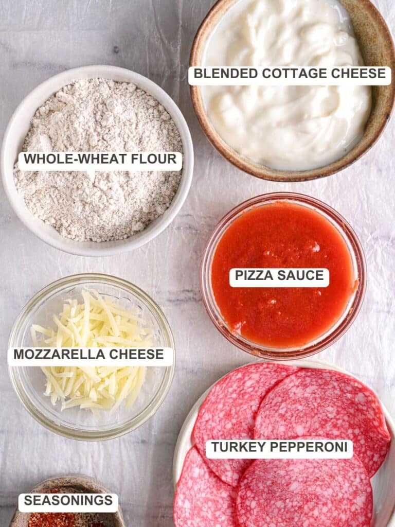 Pizza ingredients laid out – A top-down view of the key ingredients for cottage cheese pizza crust: whole wheat flour, cottage cheese, sugar-free pizza sauce, shredded cheese, and turkey salami.