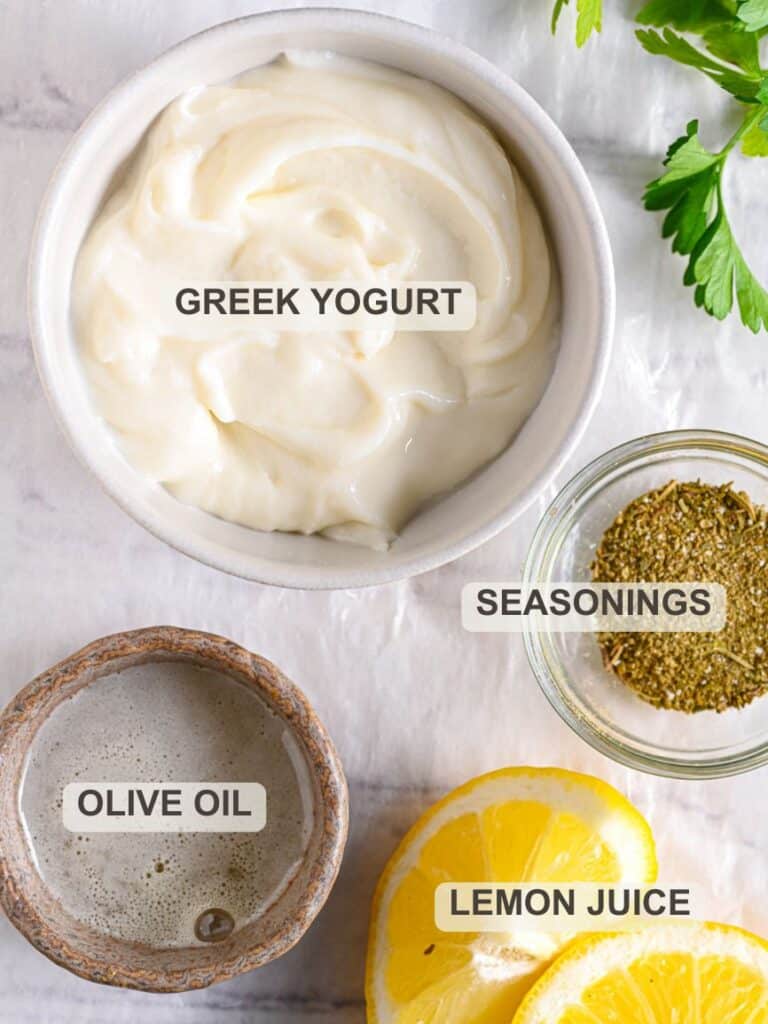 Homemade healthy Greek yogurt ranch dressing with ingredients like lemon, seasonings, herbs, and olive oil on a white surface.