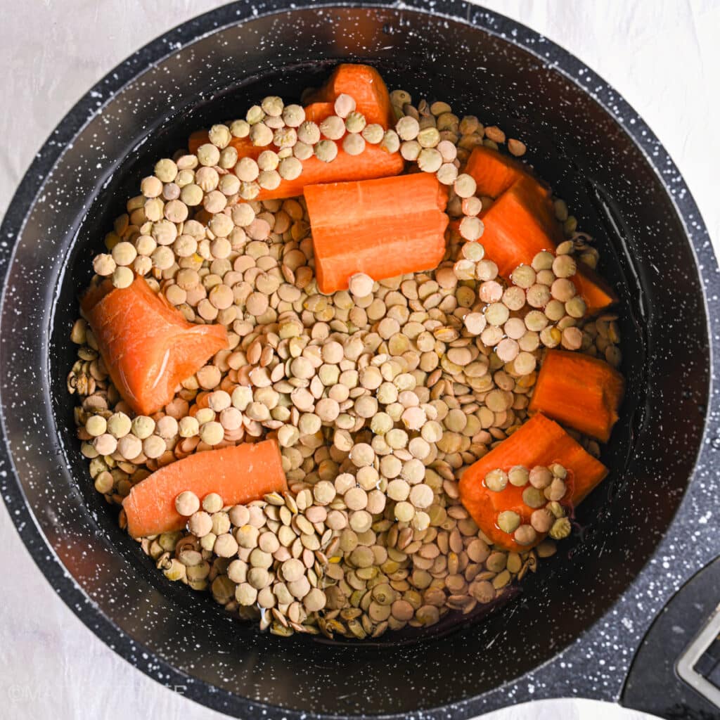 Uncooked French lentils and chopped carrots simmering in a black pot for lentil salad
