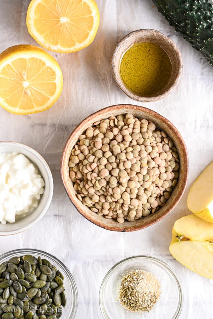 Lentil salad ingredients laid out on table including dry French lentils, lemon, olive oil, apple, pumpkin seeds, and cottage cheese