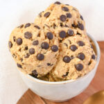 A white bowl filled with scoops of chocolate chip protein cookie dough, placed on a wooden surface with a white cloth in the background.