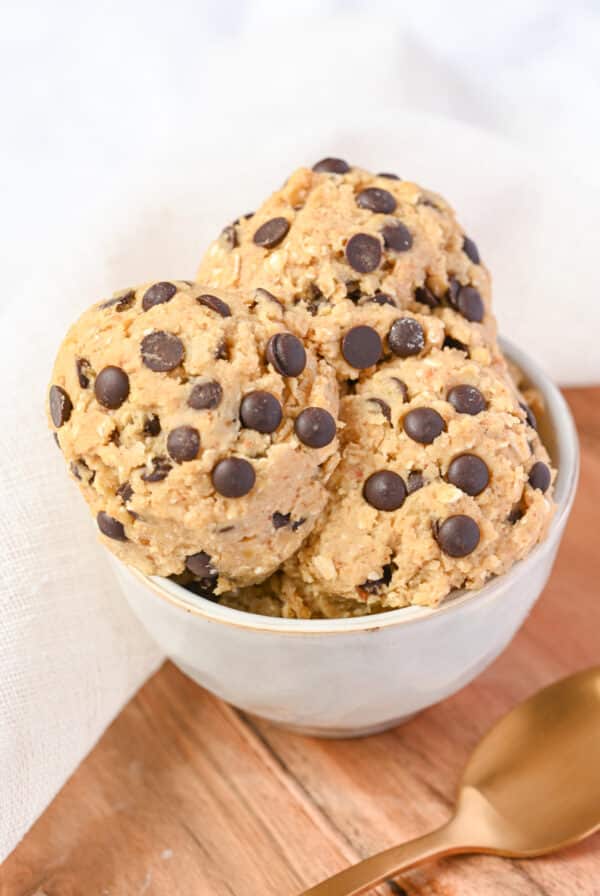 A white bowl filled with scoops of chocolate chip protein cookie dough, placed on a wooden surface with a white cloth in the background.