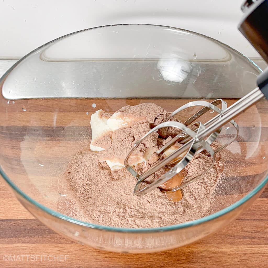 Hand mixer positioned over yogurt and protein powder, ready to whip into chocolate protein mousse.