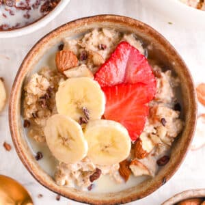 Bowl of Vanilla Protein Oatmeal with sliced Banana, Strawberries and Cocoa Nibs surrounded by Chocolate Protein Oatmeal and Protein Powder Apple Cinnamon Porridge.