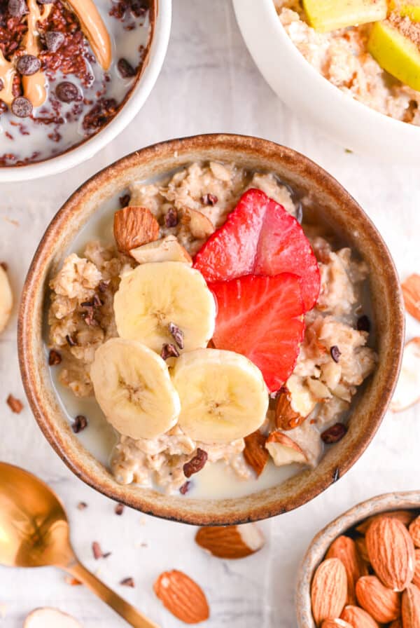 Bowl of Vanilla Protein Oatmeal with sliced Banana, Strawberries and Cocoa Nibs surrounded by Chocolate Protein Oatmeal and Protein Powder Apple Cinnamon Porridge.