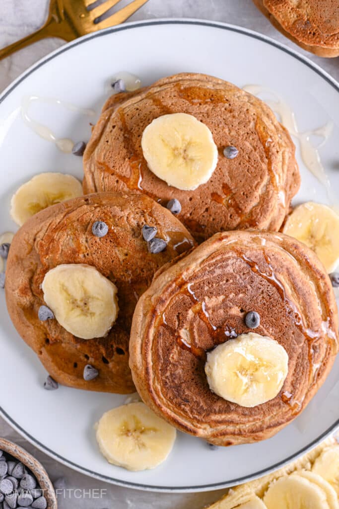 Fluffy egg white pancakes served on a white plate with sliced banana, dark chocolate chips and a drizzle of maple syrup.