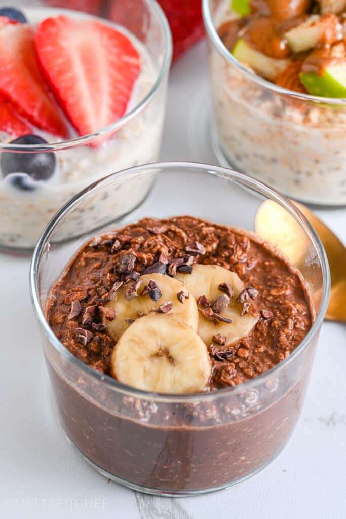 Chocolate overnight oatmeal topped with sliced banana and cocoa nibs; with other overnight oats flavors in the background