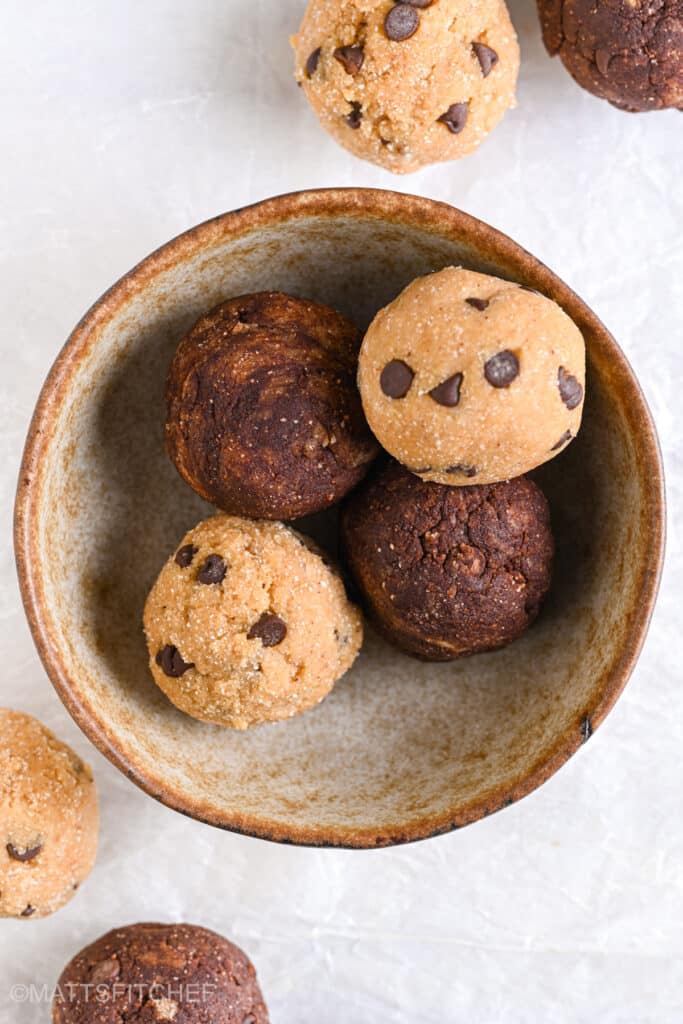 A ceramic bowl filled with chocolate chip protein balls, featuring a mix of cocoa and classic dough bites, placed on a light-colored surface.