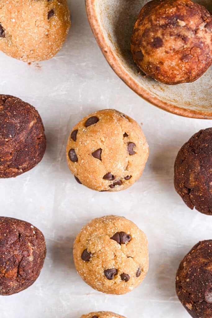 Chocolate chip protein balls, both cocoa-coated and uncoated, scattered on a neutral background, with a ceramic bowl partially visible.