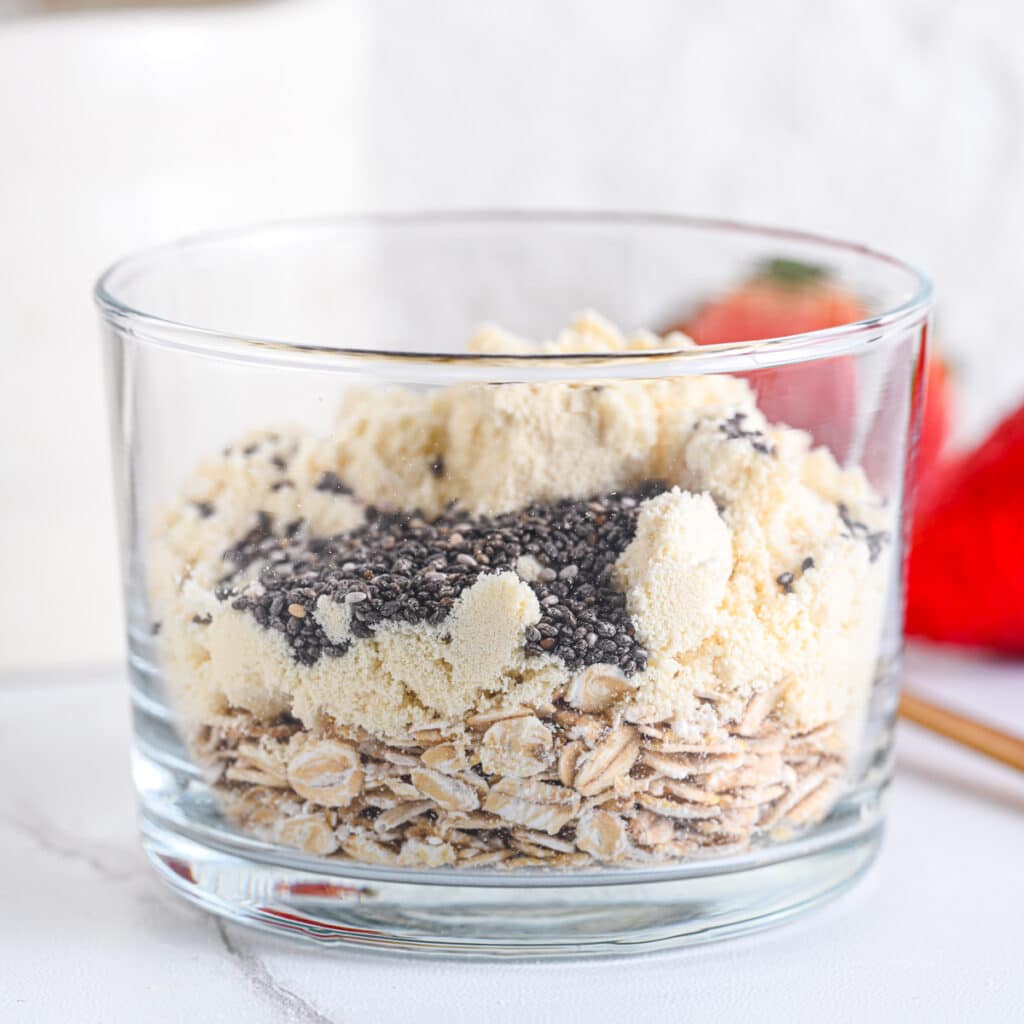 Chia seeds, protein powder and old-fashioned rolled oats in a glass, ready to stir in creamy protein overnight oats