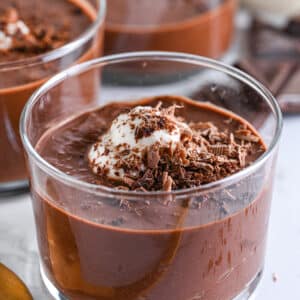 Sugar-free chocolate pudding from close, topped with unsweetened Greek yogurt and sugar-free shaved chocolate. Other 2 glasses with pudding are to be seen in the background, together with pieces of dark chocolate and a golden spoon ready to serve.