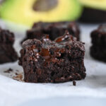 Close-up of a fudgy chocolate avocado brownie with ripe avocados shown in the background.