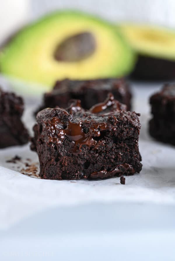 Close-up of a fudgy chocolate avocado brownie with ripe avocados shown in the background.
