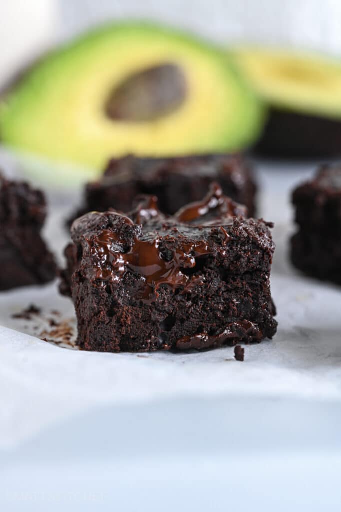 Close-up of a fudgy chocolate avocado brownie with ripe avocados shown in the background.