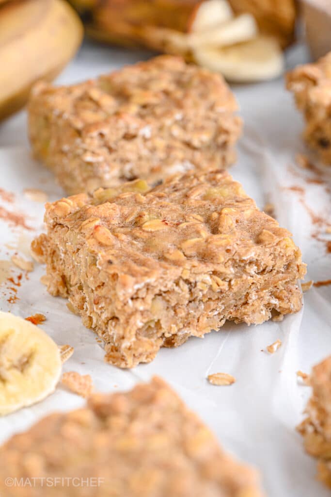 3 sliced banana oat bars cut in squares, with ripe bananas and flakes of oats in the background