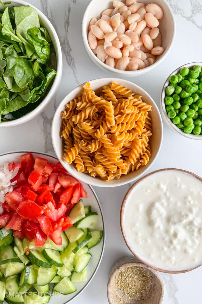 Ingredients for high protein pasta salad &ndash; chickpea rotini noodles, cottage cheese, white beans, peas, chopped spinach, diced tomatoes, sliced cucumber and sliced onion plus seasonings