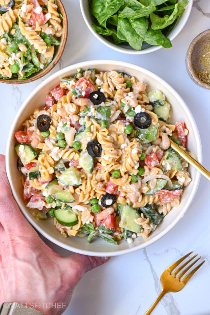 Left hand of Matteo, the fitness chef and cook of this recipe, holding a plate loaded with healthy high protein pasta salad. Perfect as a lunch or dinner that does not require cooking
