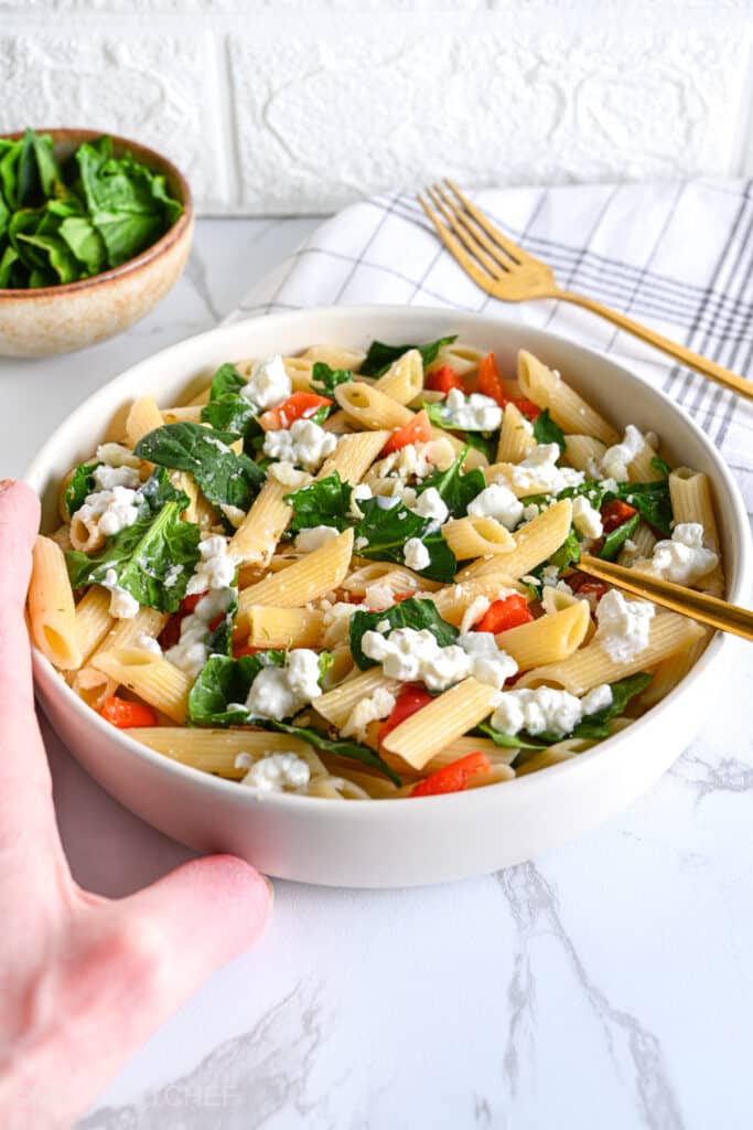 Low calorie pasta salad shown from the side, with creamy cottage cheese for protein and vibrant fresh vegetables for fiber and nutrients. On the left side, the hand of the fitness chef and fat loss coach can be seen, ready to dig in and enjoy a forkful.