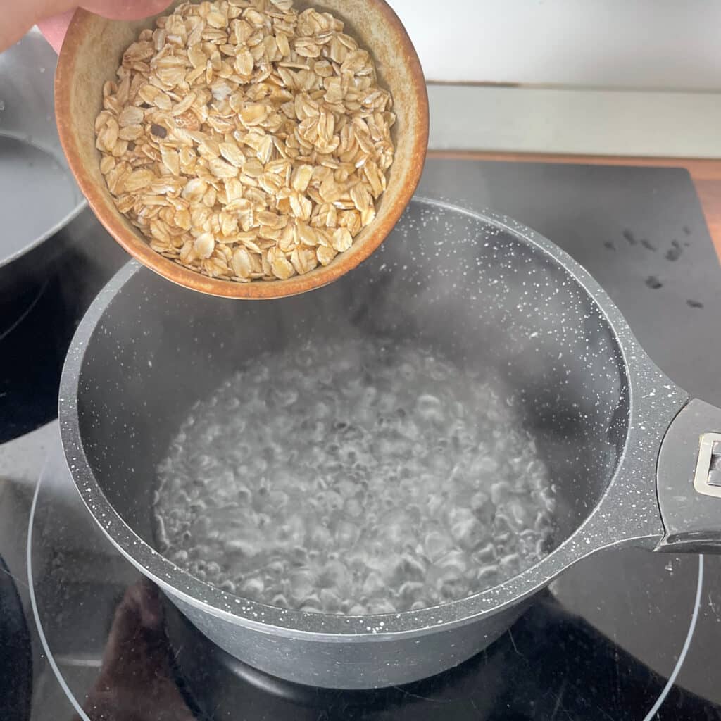 Rolled oats being added with a rustic bowl to a pot of boiling water for oatmeal